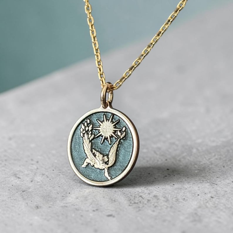 May include: Gold-toned necklace featuring a circular pendant with a teal background. The pendant showcases a sunburst design with a bird-like figure and wheat stalks. The necklace is a delicate piece of jewelry.