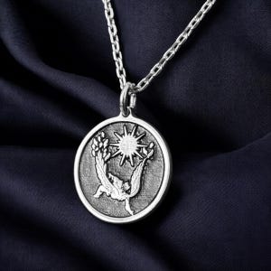 May include: Silver-toned pendant necklace featuring a circular pendant with a detailed relief design. The pendant depicts a stylized sun, a bird, and foliage. The necklace chain is also silver-toned, and the background is a dark blue fabric.
