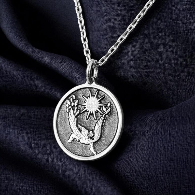 May include: Silver-toned pendant necklace featuring a circular pendant with a detailed relief design. The pendant depicts a stylized sun, a bird, and foliage. The necklace chain is also silver-toned, and the background is a dark blue fabric.