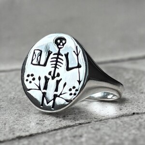 May include: A silver signet ring with a black and white engraved design of a skeleton holding a trident. The ring is oval shaped and has a smooth, polished finish.