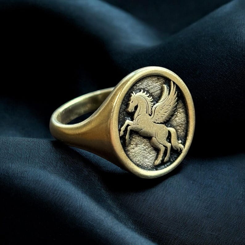 May include: A gold-colored ring with a detailed Pegasus design. The ring features a round face with the winged horse in relief. The band and face have a slightly aged appearance. The ring is set against a dark, textured background.
