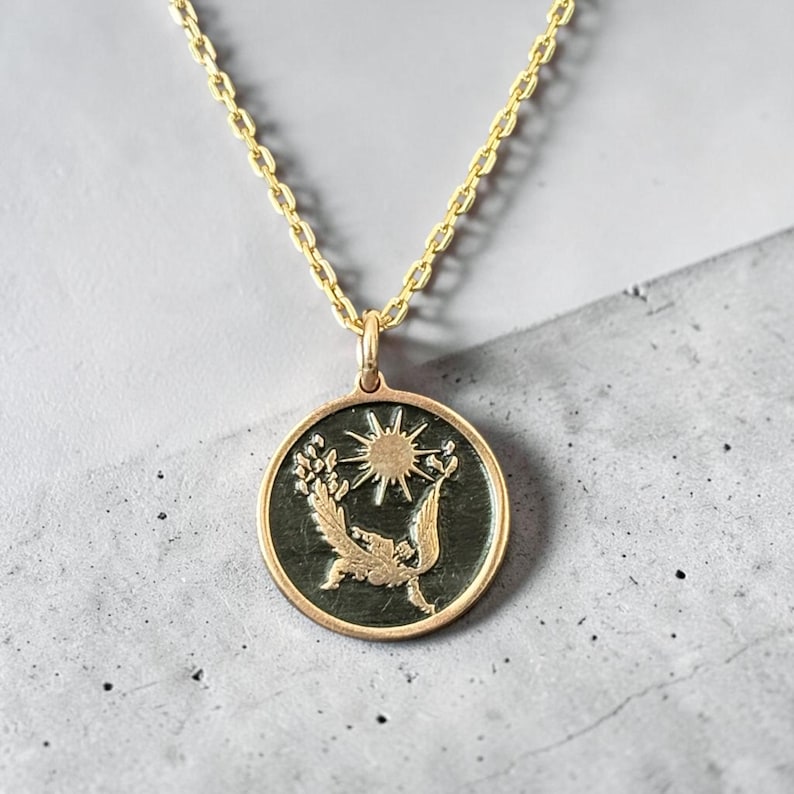 May include: Gold-toned pendant necklace featuring a circular charm with a sun and floral design. The pendant hangs from a delicate chain. The charm has a dark background with gold-colored details. The necklace is displayed on a gray surface.