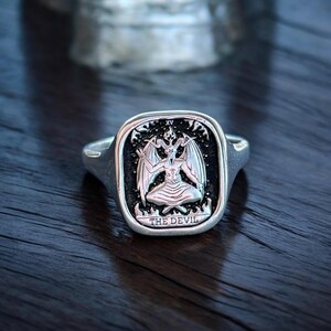 K&ouml;nnte beinhalten: Ein silberner Ring mit einer schwarzen quadratischen Fassung, die eine Darstellung der Tarotkarte des Teufels zeigt. Die Karte zeigt eine geh&ouml;rnte Gestalt mit Fl&uuml;geln, die auf einem Thron mit Flammen an der Basis sitzt. Der Text "THE DEVIL" ist unter der Figur eingraviert.