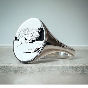May include: A silver signet ring with an oval shaped face featuring an engraved image of a couple embracing.