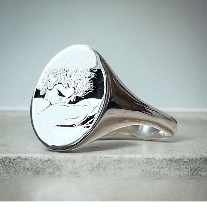 May include: A silver signet ring with an oval shaped face featuring an engraved image of a couple embracing.