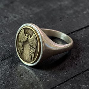 May include: Silver signet ring with an oval face featuring a detailed bronze-colored wing design. The ring has a polished finish and a classic, minimalist style. The wings are intricately detailed, adding a touch of elegance to the jewelry.