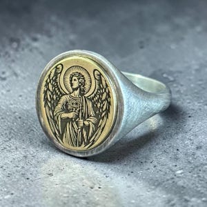 May include: Silver signet ring with an oval gold-coloured face featuring an engraved image of an angel with wings, holding a staff. The ring has a brushed metal finish and a classic design.