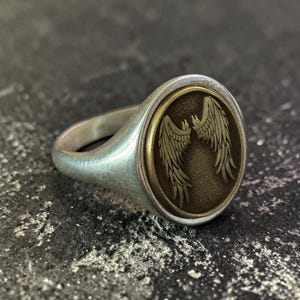 May include: Silver signet ring with an oval face. The face features a detailed design of two symmetrical wings in a bronze-colored oval. The ring has a polished finish and a classic, elegant design.
