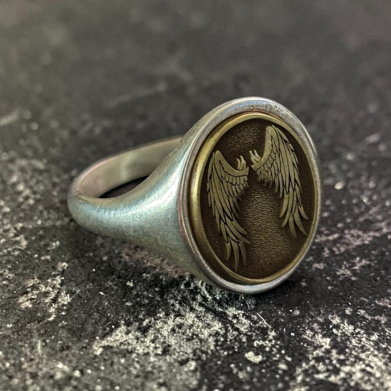 May include: Silver signet ring with an oval face. The face features a detailed design of two symmetrical wings in a bronze-colored oval. The ring has a polished finish and a classic, elegant design.