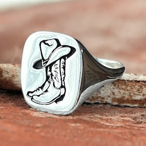 May include: A silver signet ring with a square face featuring an engraved design of a cowboy boot with a hat on top.