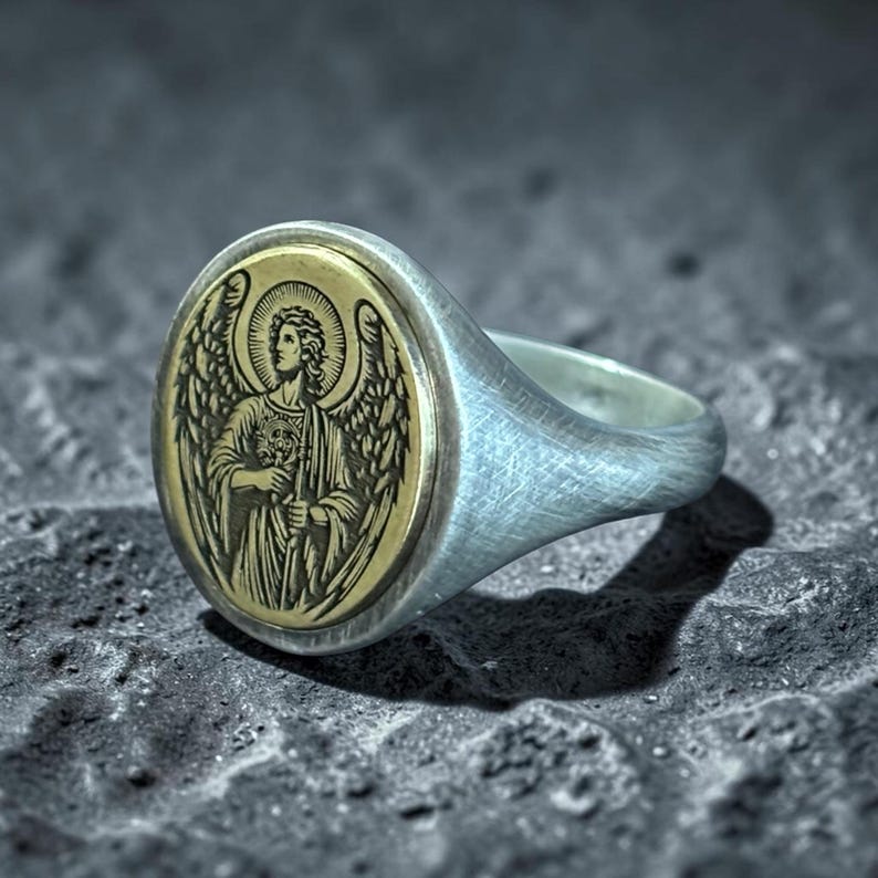May include: A silver ring with an oval, gold-coloured face featuring an engraved angel with outstretched wings. The ring has a brushed metal finish and is set against a dark, textured background.