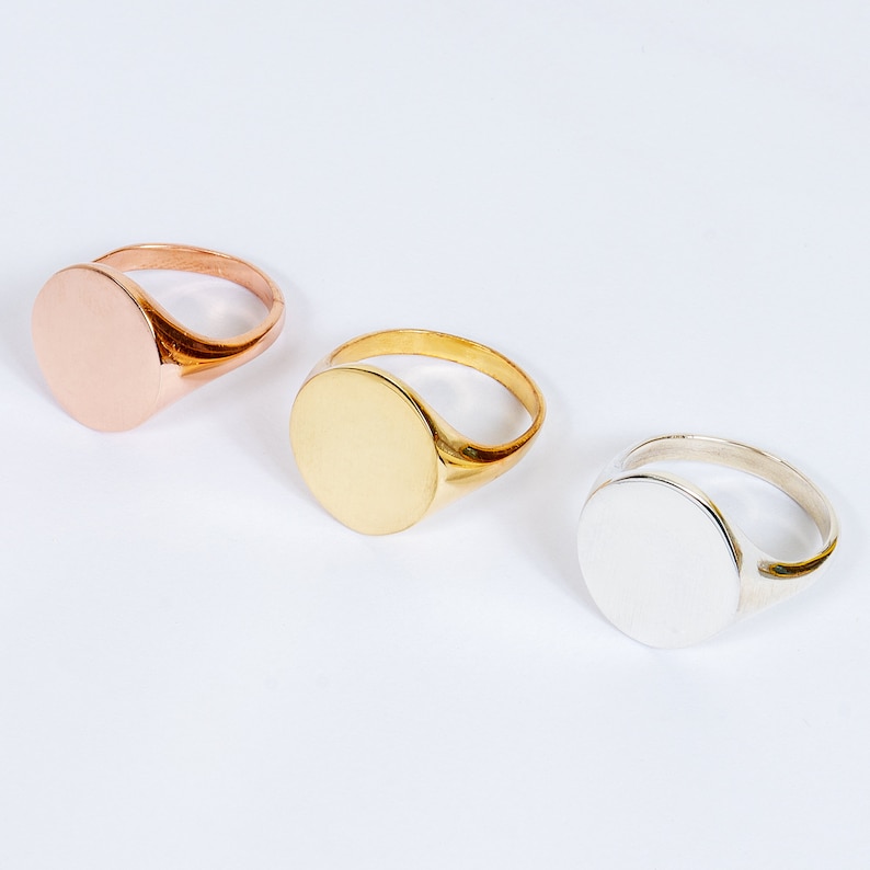 May include: Three signet rings in rose gold, gold, and silver. Each ring features a smooth, circular face and a simple band. The rings are displayed on a white surface, showcasing their polished finish and varying metallic tones.