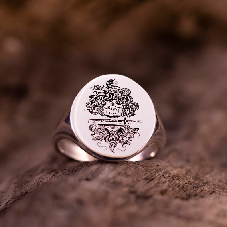 May include: A silver signet ring with a black and white engraved image of Medusa, a mythical Greek figure with snakes for hair, and a sword through her head.