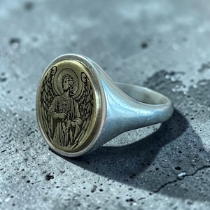 May include: Silver signet ring with an oval gold-toned face featuring an engraved angel design. The angel has detailed wings and a halo. The ring has a smooth, polished finish.