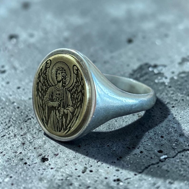 May include: Silver signet ring with an oval gold-toned face featuring an engraved angel design. The angel has detailed wings and a halo. The ring has a smooth, polished finish.