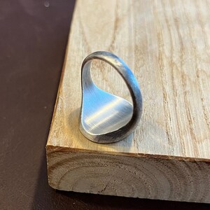 May include: A silver signet ring with a smooth, rounded surface. The ring is shown from the side, with the band facing the viewer.