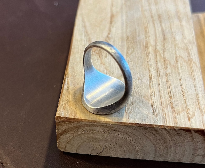 May include: A silver signet ring with a smooth, rounded surface. The ring is shown from the side, with the band facing the viewer.