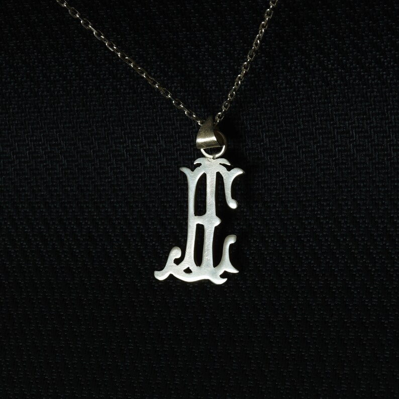 May include: A silver chain necklace with a pendant in the shape of the letter "E". The pendant is ornate and has a decorative design.