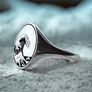 May include: A silver ring with an oval-shaped top featuring a black and white image of a large wave crashing against a mountain.