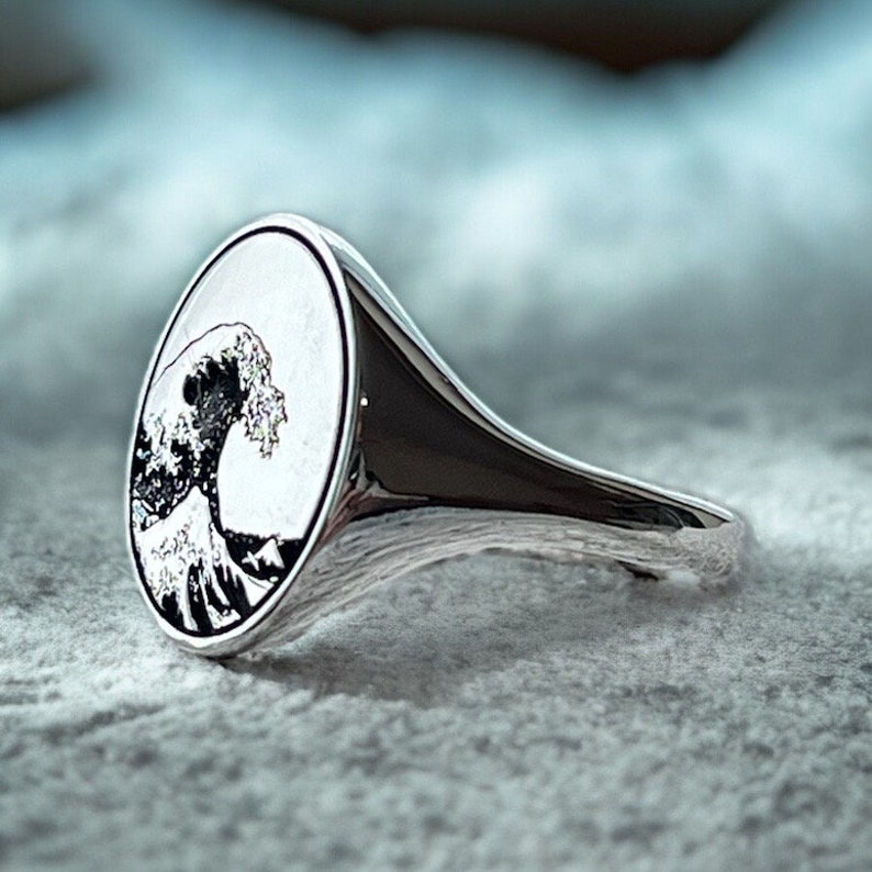 May include: A silver ring with an oval-shaped top featuring a black and white image of a large wave crashing against a mountain.