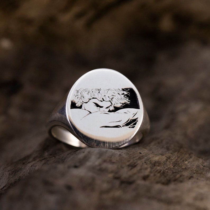 May include: A silver signet ring with an engraved image of a couple embracing. The ring is oval shaped and has a smooth, polished finish.