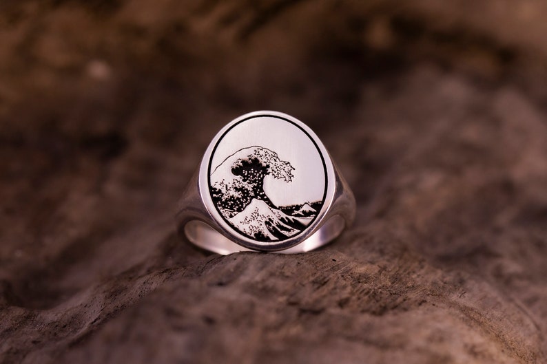 May include: A silver ring with an oval-shaped design featuring a black and white engraved image of a large wave.