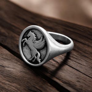 May include: Silver signet ring featuring a detailed relief of a winged horse, or Pegasus, within an oval frame. The ring has a polished finish and a thick band. The design is set against a dark wooden surface.
