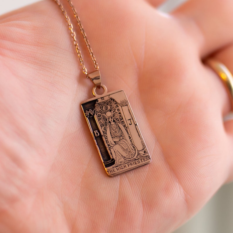 Op de afbeelding: Een gouden ketting met een hanger met de tarotkaart van de Hogepriesteres. De hanger is rechthoekig en heeft een gedetailleerde gravure van de afbeeldingen op de kaart.