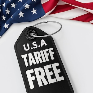 以下が含まれることがあります： 白い文字で「U.S.A TARIFF FREE」と書かれた黒いタグ。銀色のループが付いており、背景にはアメリカ国旗があります。星が散りばめられた青い部分と、赤と白のストライプが見えます。