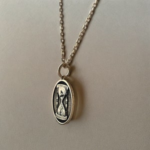 Pu&ograve; includere: Una collana con catena d'argento con un ciondolo ovale che presenta un design a clessidra e l'iscrizione "Memento Mori" attorno al bordo.