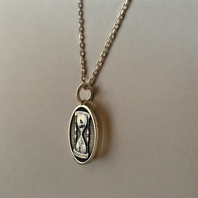 Pu&ograve; includere: Una collana con catena d'argento con un ciondolo ovale che presenta un design a clessidra e l'iscrizione "Memento Mori" attorno al bordo.