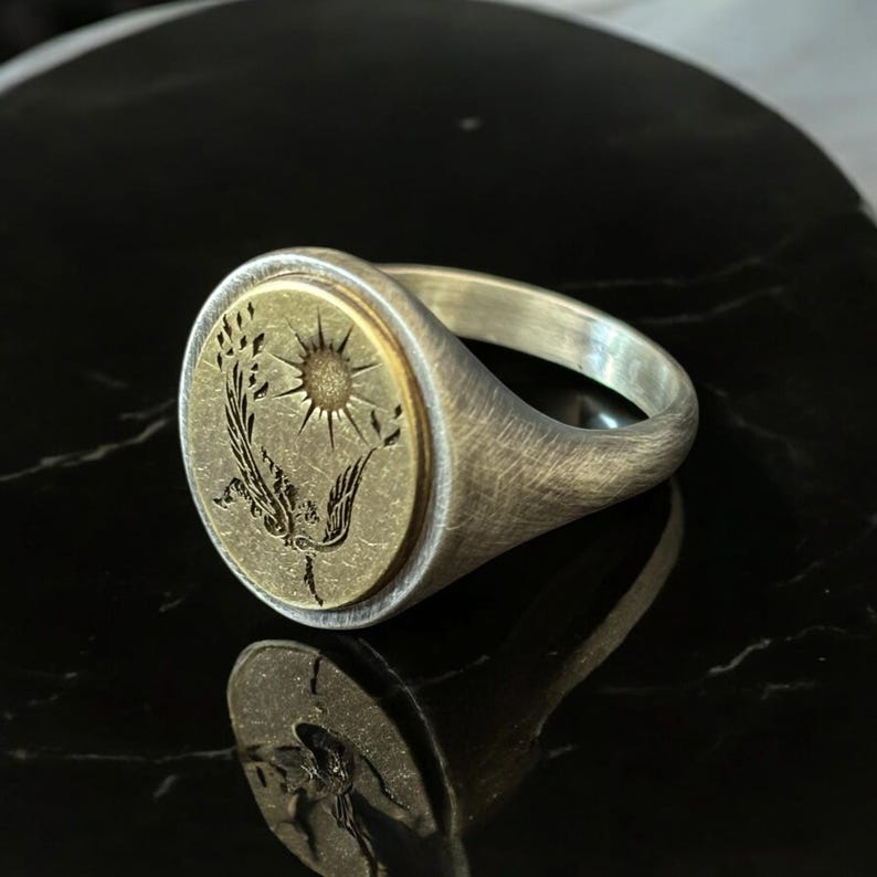 May include: A silver signet ring with a brushed finish. The ring features a circular gold-toned face with an engraved design of a sun, a bird, and foliage. The ring is set against a black, reflective surface.