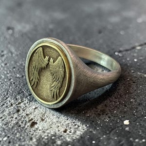 May include: A silver ring with a brushed finish. The ring features a circular emblem with a gold-colored border, showcasing a detailed design of stylized wings. The ring is a piece of jewelry.