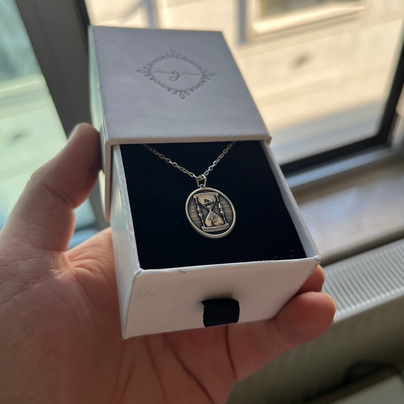 Pu&ograve; includere: Una collana d'argento con un ciondolo a forma di clessidra. Il ciondolo &egrave; di forma ovale e presenta un design inciso di una clessidra con le parole "Il tempo non aspetta nessuno" incise su di esso.