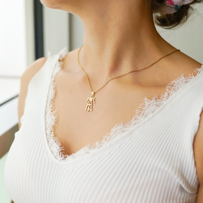 May include: A gold necklace with a delicate chain and a pendant featuring a stylized monogram. The necklace is worn by a person with a white tank top.