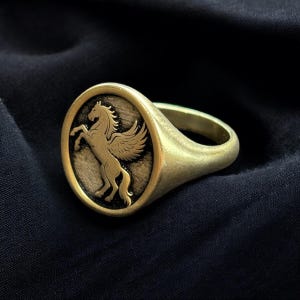 May include: A gold-colored ring with an oval face featuring a detailed relief of a winged horse, likely a Pegasus. The ring has a smooth, rounded band and a slightly aged appearance, suggesting a vintage or antique style.