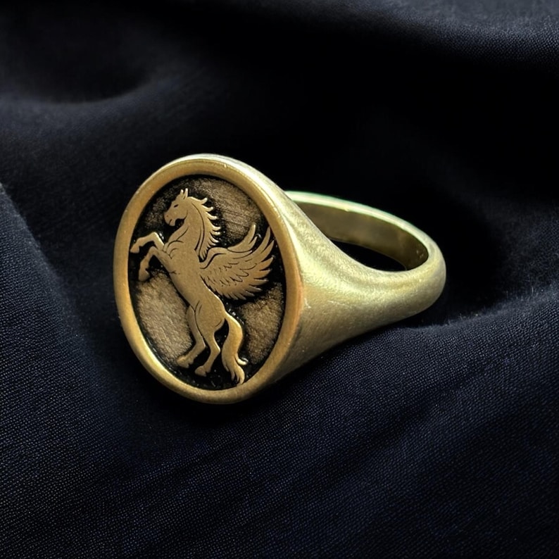May include: A gold-colored ring with an oval face featuring a detailed relief of a winged horse, likely a Pegasus. The ring has a smooth, rounded band and a slightly aged appearance, suggesting a vintage or antique style.