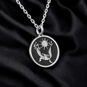 May include: Silver-toned pendant necklace featuring a circular pendant with a detailed design. The pendant has a black background with a sunburst and a winged figure. The chain is a delicate link style.