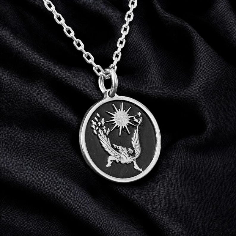 May include: Silver-toned pendant necklace featuring a circular pendant with a detailed design. The pendant has a black background with a sunburst and a winged figure. The chain is a delicate link style.