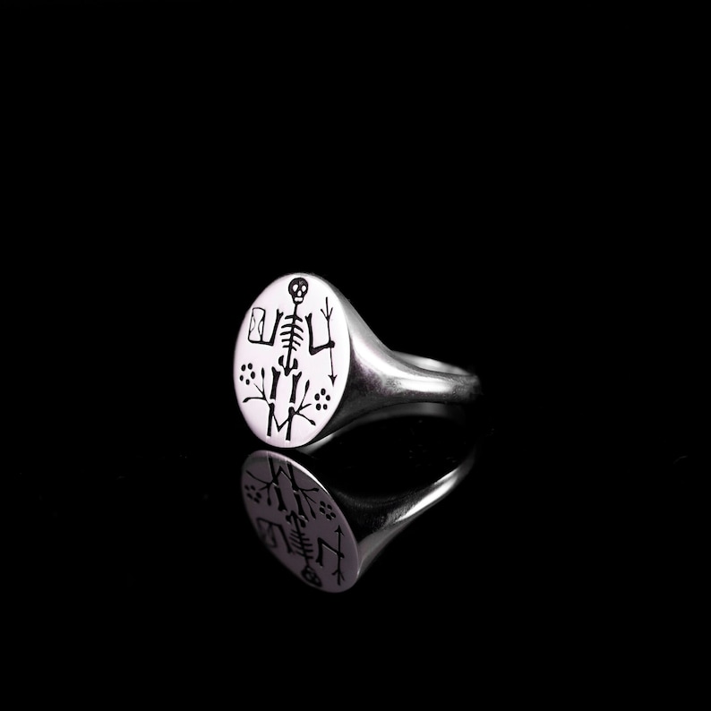 May include: A silver signet ring with a round, black and white engraved design of a skeleton with a skull. The ring is on a black background.