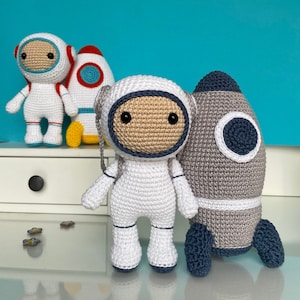 Può includere: Bambola astronauta all'uncinetto, tuta spaziale bianca, viso beige e bordi blu scuro. Un razzo grigio con un cerchio bianco e blu scuro è accanto all'astronauta. Altre bambole e un razzo sullo sfondo. Le bambole sono fatte a mano.