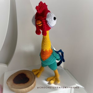 May include: Crocheted rooster toy with a red comb, yellow and orange neck, teal body, and large white eyes with black pupils. The rooster is standing on a white surface with a wooden slice behind it. The text "@CROCHETOXENTEMYFRIEND" is visible in the bottom right corner of the image.