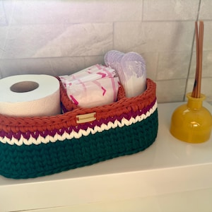 May include: A handmade, oval-shaped storage basket in green, orange, white, and purple. The basket holds a roll of toilet paper and packaged sanitary products. A yellow diffuser bottle with reeds sits beside the basket.