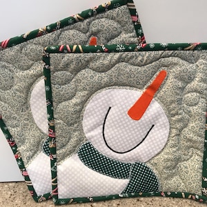 May include: Two quilted pot holders with a snowman design. The pot holders are green with a white snowman face and a green scarf. The background fabric is a light green with a floral pattern.
