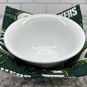 May include: A green and white fabric bowl cozy with the word "Packers" printed on it. A white ceramic bowl is sitting inside the cozy.