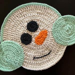 May include: A round, crocheted coaster featuring a cream-colored face with two black circular eyes, an orange carrot-shaped nose, and a dark blue stitched smile. The coaster is accented with light green earmuffs on both sides of the face.