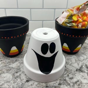 May include: Three painted terracotta pots, one white with a ghost face, two black with candy corn designs. The black pots are filled with candy corn.