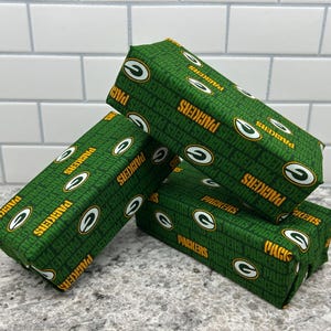 May include: Three rectangular objects are stacked, each wrapped in green fabric. The fabric is patterned with the word "PACKERS" in gold, along with the team's logo. The objects are arranged on a speckled gray surface.