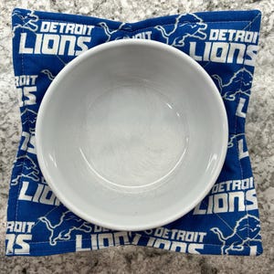 May include: A white ceramic bowl sits on a blue Detroit Lions themed bowl cozy. The cozy is quilted and features the team's logo and name in white. The background is a light gray.