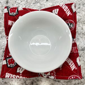 May include: A white ceramic bowl sits on a red and white fabric bowl cozy. The cozy features a repeating pattern of the letter "W" and the word "WISCONSIN" in white. The bowl is centered on the square cozy.